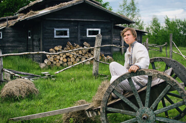 young man in national costume sits in an old cart against the background of a village house