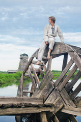 young man in a national costume of white linen sits on an old water mill wheel and looks into the distance
