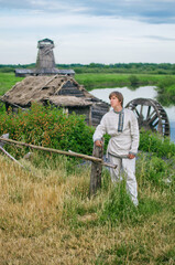 young man in national costume stands near a water mill near an ancient settlement
