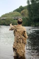 man in rags stands in the shallows of the river reaching for an old wooden bridge
