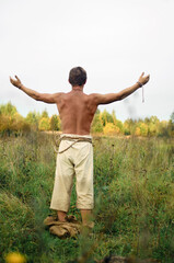 young man stripped to the waist stands in a wild field with his arms outstretched