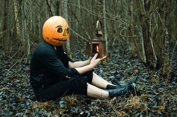 man in ragged clothes with a lantern and a Halloween mask on his head is sitting in a damp and gloomy forest on the ground. pumpkin Jack