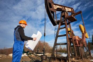 Side view of oil worker holding plan of oil field. Engineer in work uniform and helmet standing near petroleum pump jack under beautiful cloudy sky. Concept of petroleum industry and oil extraction.