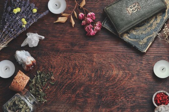 Dark brown wooden table background frame for text, decorated with witchy items to look esoteric and occult. Free writing space in the middle. Dried herbs, crystals, old vintage books candles flowers