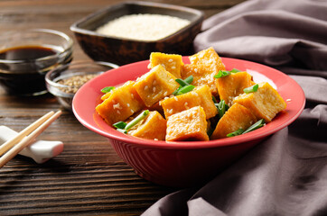 Crispy stir fried tofu cubes with chives in clay dish on wooden kitchen table with napkin and soy sauce aside