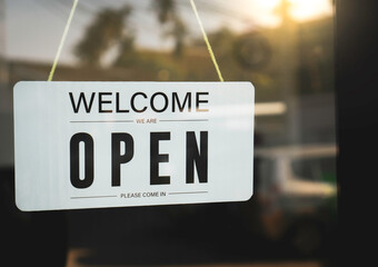 The signage welcomes we are open on a white acrylic sign hanging on the glass door in front of the...