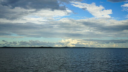 Fototapeta na wymiar Cloudscape with grey and white clouds against a blue sky fading to pale aqua near the horizon, with blue-grey sea in the foreground and land on the horizon. Moreton Bay, Queensland, Australia.