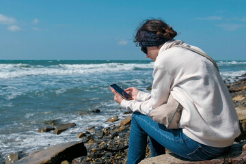 Woman is reading something on phone sitting on the sea shore in autumn