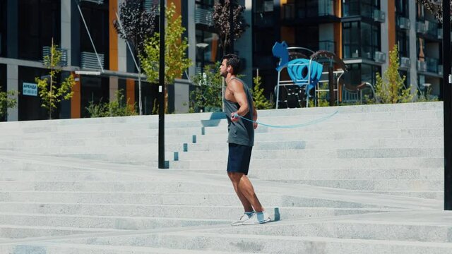 Young man exercising outside. Guy using jumping rope for exercising. Training on street alone during sunny warm day. Powerful slim and well-built person doing fitness.