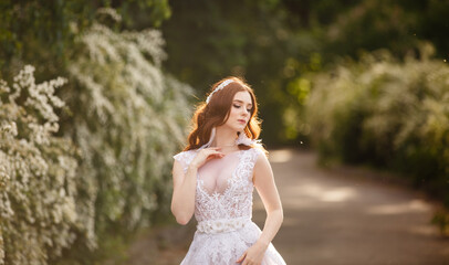 Fototapeta na wymiar Beautiful redhead Bride in fantastic wedding dress in blooming garden. Growth Portrait in sunset light. Pretty young caucasian redhead girl walks in a garden and plays with her dress. Young princess