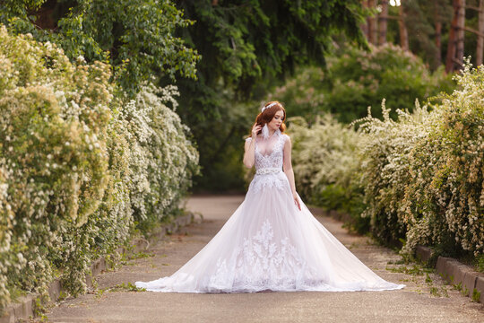 Beautiful redhead Bride in fantastic wedding dress in blooming garden. Growth Portrait in sunset light. Pretty young caucasian redhead girl walks in a garden and plays with her dress. Young princess