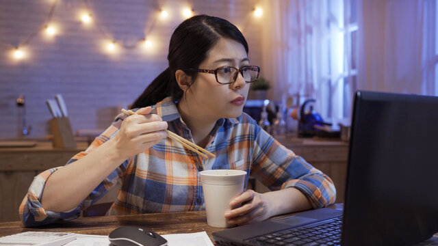 Young Asian Chinese Woman Eating Instant Noodles At Home Workplace Being Too Busy With Work. Starving Girl Holding Chopsticks With Ramen Soup In Cup And Staring At Laptop Computer Screen In Evening.