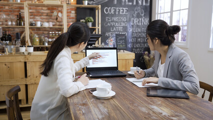 side view of two happy businesswomen sharing financial report on laptop together. asian korean female coworkers discussing chart on notebook computer in cafe store. professional women colleagues