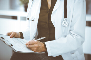 Unknown woman-doctor writing something at clipboard while sitting at the chair, close-up. Therapist wearing green blouse at work is filling up medication history record. Medicine concept