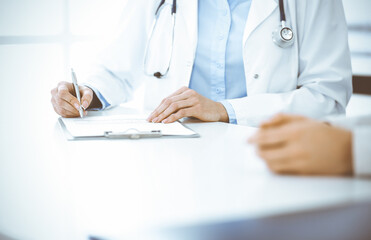 Unknown woman-doctor and female patient sitting and talking at medical examination in clinic, close-up. Therapist wearing blue blouse is filling up medication history record. Medicine concept