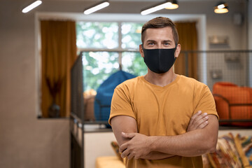 Social distancing at work. Portrait of young confident man, male office worker in protective face mask keeping arms crossed, looking at camera