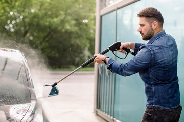 Handsome bearded young man in jeans shirt, washing his new car manually with water high-pressure hose at outdoor self wash service. Car washing concept.