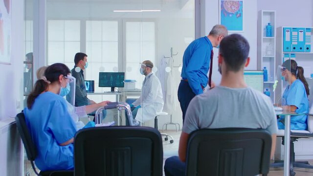 Doctor with visor against coronavirus and patient in examination room. Senior man with face mask talking with nurse at reception. Assistant and disabled woman with walking frame in waiting area.