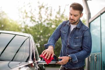 Portrait of handsome Caucasian bearded man in jeans shirt, wiping side mirror with red microfiber rag at outdoor car wash self station.