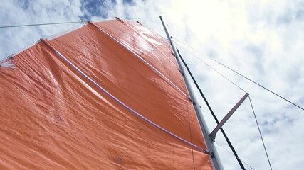 Mast and mainsail of a sailboat with ropes against the white clouds, view from below. Orange mainsail and metal mast. Shot from lower anlge on sunny day.