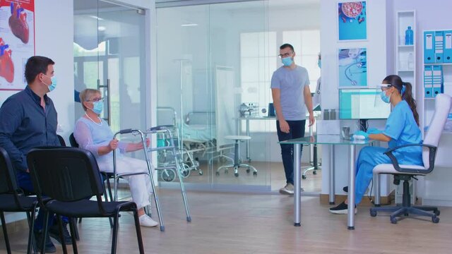 Invalid senior woman with walking frame wearing face mask against coronavirus, discussing with nurse in hospital waiting area. Senior man at clinic reception. Health care system duting global pademic