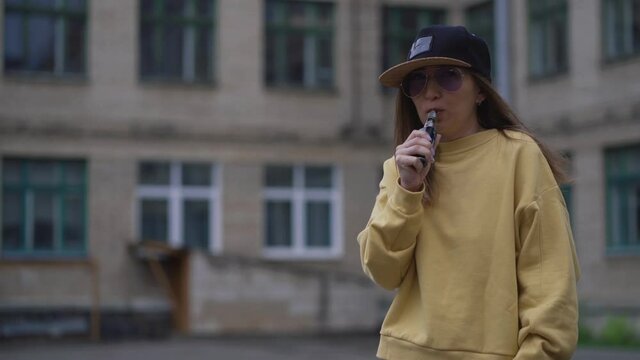 Pretty young hipster woman in black cap yellow hoodie vape ecig. Girl vape popular ecig gadget, vaping device.Happy brunette vaper girl with e-cig. Portrait of smoker female model with electronic