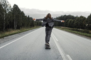 Man in grey office suit with long blond hairs is riding skateboard longboard down road outside the city, back view. Freedom from office work concept. He is riding hands up and enjoying his trip.