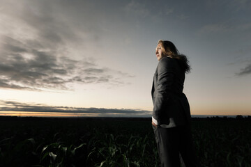 Fototapeta premium young businessman stands in a field in a gray suit at sunset. Blond with long hair in a cornfield