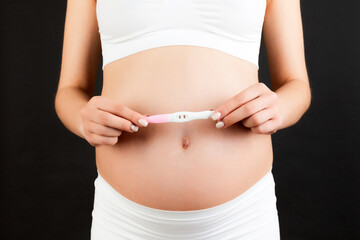 Close up portrait of pregnant woman in white underwear holding positive pregnancy test against her belly at black background. Healthy pregnancy. Copy space