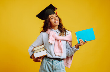 Graduate girl in a graduation hat on her head, with books stands on a yellow background. African...