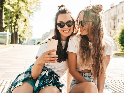 Two young smiling hipster women in summer clothes. Girls taking selfie self portrait photos on smartphone.Models sitting in the street.Female going crazy and making funny faces in sunglasses