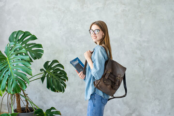 Side view of smiling girl student in glasses holding brown backpack and books isolated on grey wall background with monstera plant.Woman wearing denim clothes.Distance education in university,college. © Matveeva.photos