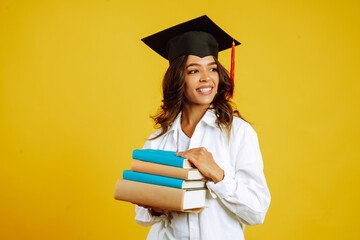 Graduate girl in a graduation hat on her head, with books stands on a yellow background. African...