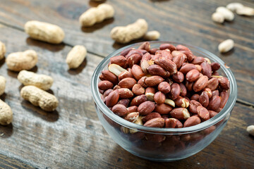 unshelled peanuts in a glass plate on a wooden background. peeled peanuts are scattered nearby. traditional beer snack. place for text.