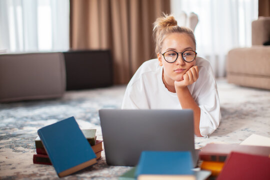 Cute thoughtful teen girl student in casual clothes, glasses lying on floor at laptop with books. Back to school