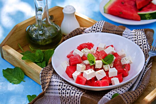 Summer salad of feta and sweet juicy watermelon in a white bowl on a wooden tray on a light blue background. Salads with watermelon.