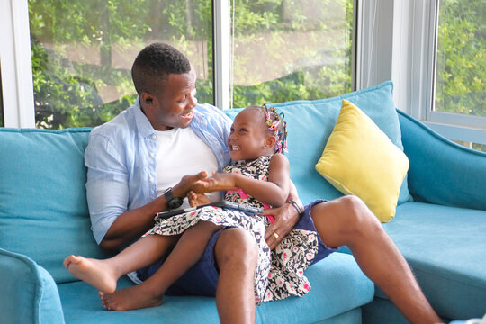 African American father playing with his little daughter who sitting on his lap while spending happy time together at home on sofa couch
