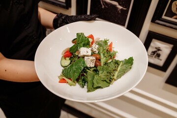 The young female waiter holding in her hands the plate with the Greek salad