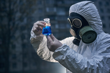 Fototapeta premium Close-up view, scientist wearing white coverall and a gas mask, holding glass flask with samples, worker observing reaction on the light, high building on background
