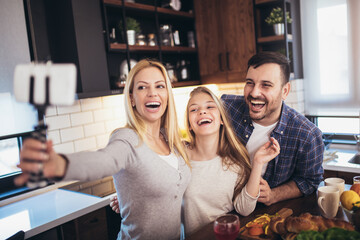 Cute girl and her beautiful parents are doing selfie using a smart phone and smiling while baking in kitchen at home