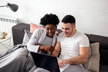 Multiethnic couple lin bed using laptop early in the morning