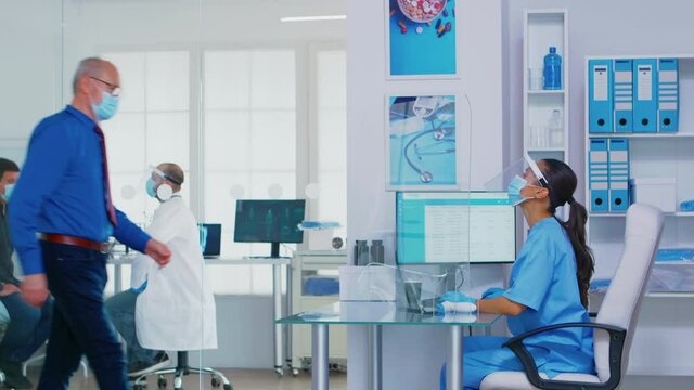 Nurse with visor against coronavirus greeting senior woman at hospital reception. Assistant with sterile gloves pushing disabled senior woman in wheelchair through waiting area.