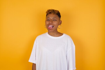 Young African American girl with afro short hair wearing white tshirt standing over isolated yellow wall showing grimace face crossing her eyes and showing tongue . Being funny and crazy