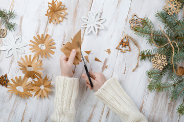 Child makes Christmas craft snowflakes from paper on wooden background. Handmade christmas gift. Top view. Christmas eve preparation. Kid's hands paking present box. Natural rustic. DIY snowflake cut