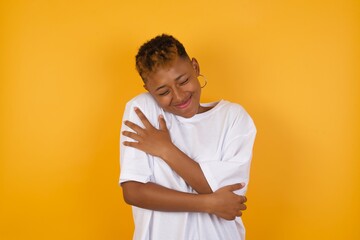 Young African American girl with afro short hair wearing white tshirt standing over isolated yellow wall Hugging oneself happy and positive, smiling confident. Self love and self care