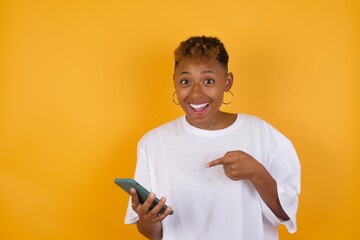 Young African American girl with afro short hair wearing white tshirt standing over isolated yellow wall holding in hands showing new cell,