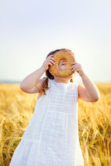 A smiling little girl in a white sundress and two pigtails on a wheat field with a bagel in the open air. Looks through the hole in the bagel with one eye.