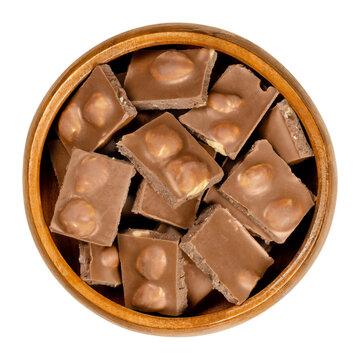 Hazelnut milk chocolate, bars broken into pieces, in wooden bowl. Small segments of candy bar. Milk chocolate with whole roasted hazelnuts, ready-to-eat. Sweet dessert. Close-up from above food photo.