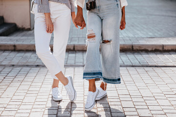 Glamorous girl in white gumshoes standing with legs crossed. Outdoor photo of two stylish ladies...