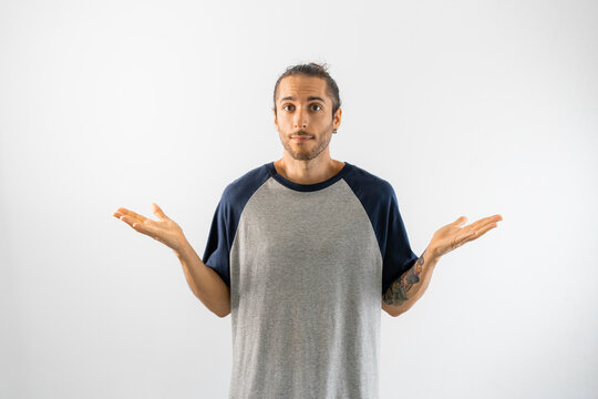 Hombre dudoso con camiseta gris de mangas azules hace el gesto de la duda en fondo blanco
